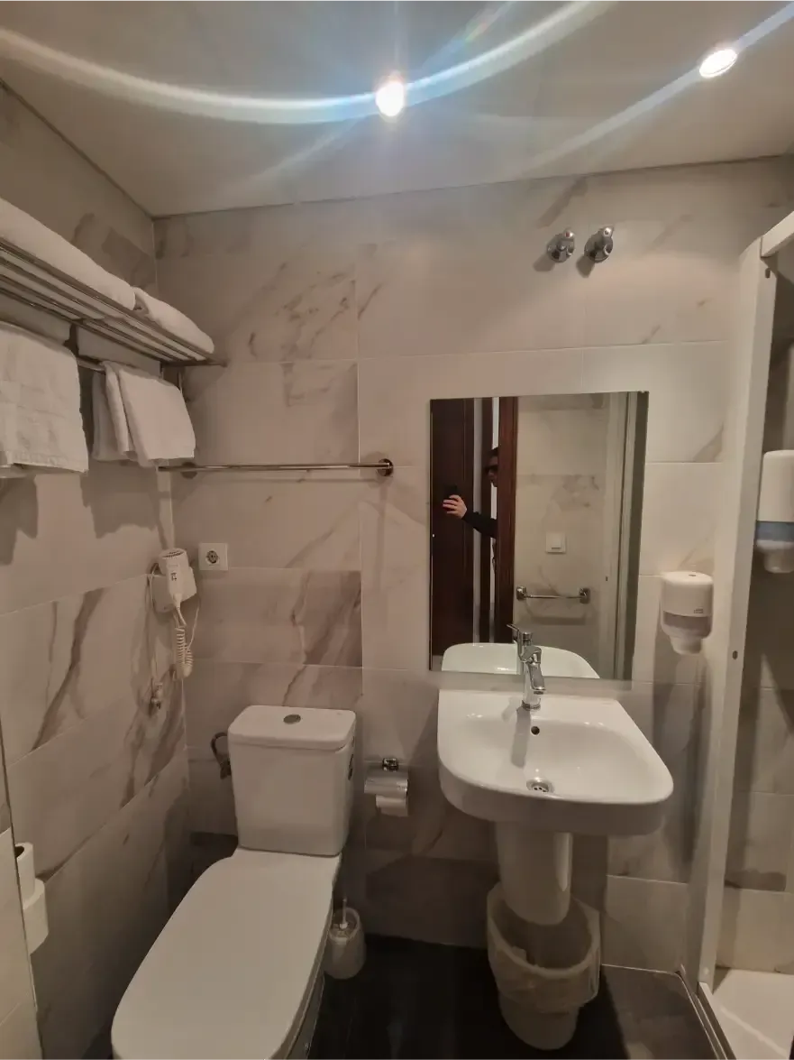 Baño pequeño con azulejos, inodoro, lavabo con pedestal, espejo y zona de ducha con iluminación bajo el techo.