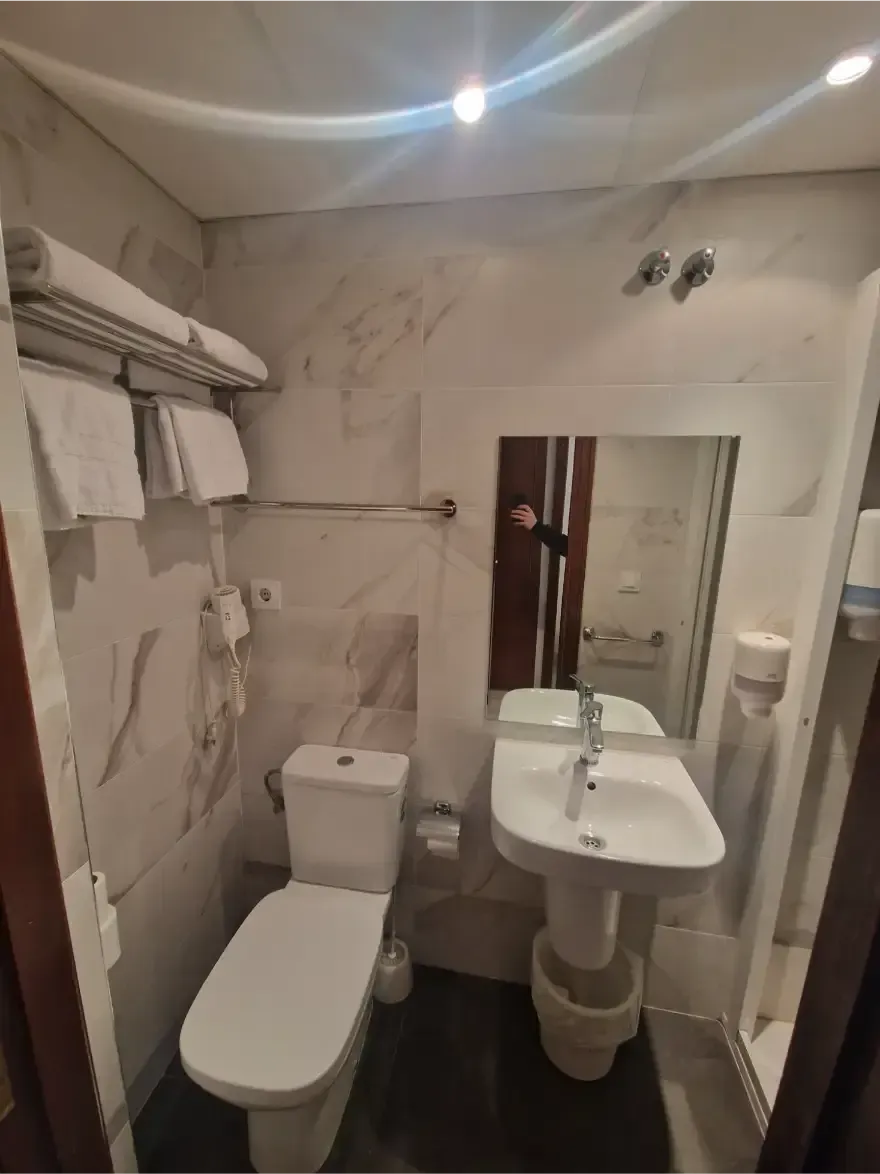 Baño pequeño con inodoro, lavabo, espejo y ducha; paredes alicatadas en blanco y luz en el techo.