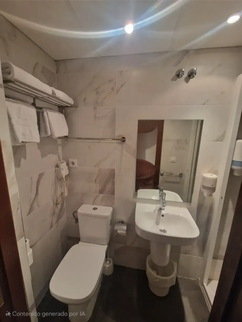 Baño pequeño con inodoro, lavabo con pedestal, espejo y toallero bajo una iluminación brillante en el techo.