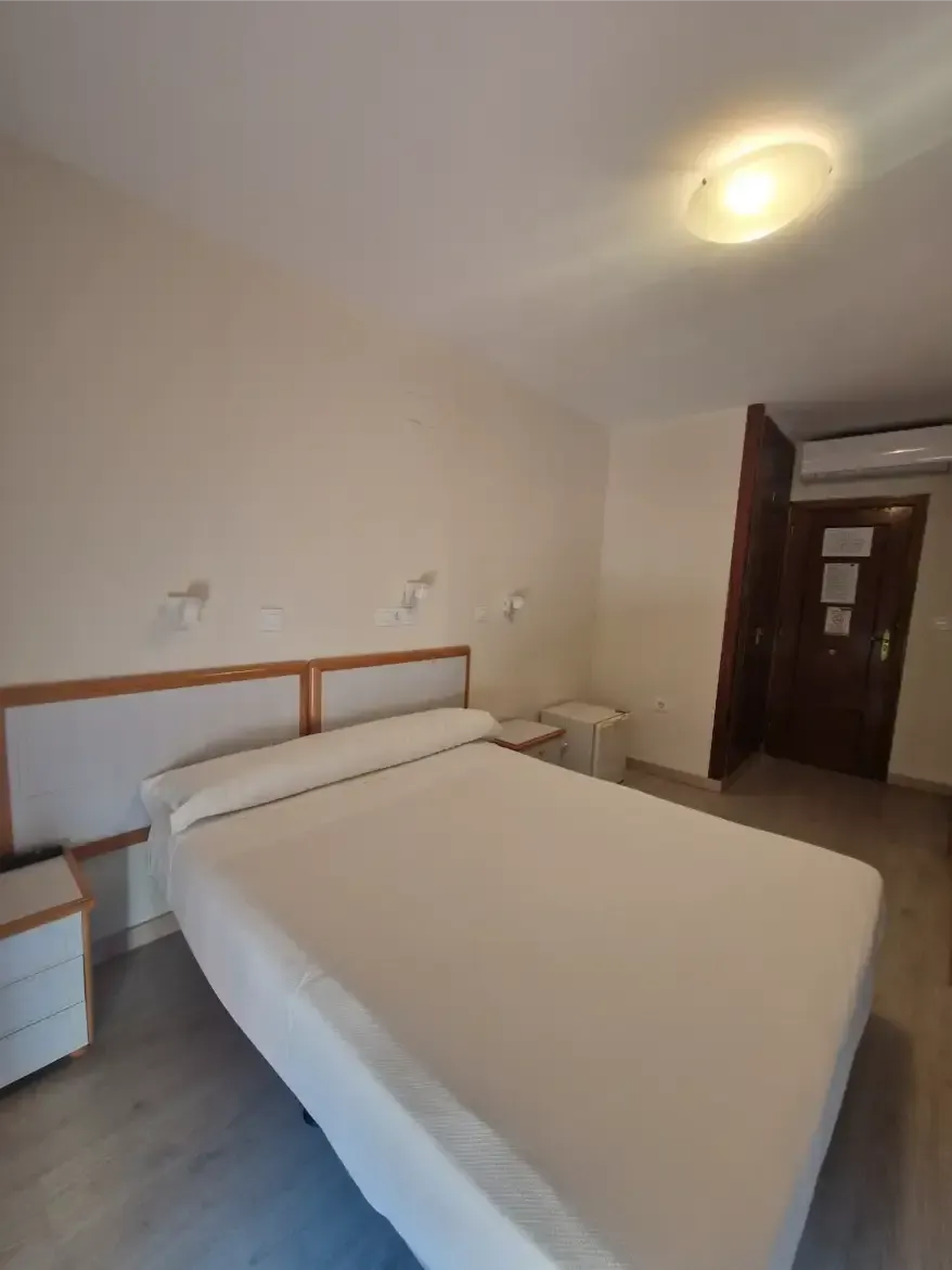 Habitación pequeña con una cama bien hecha, paredes blancas, mesita de noche y una puerta al fondo.