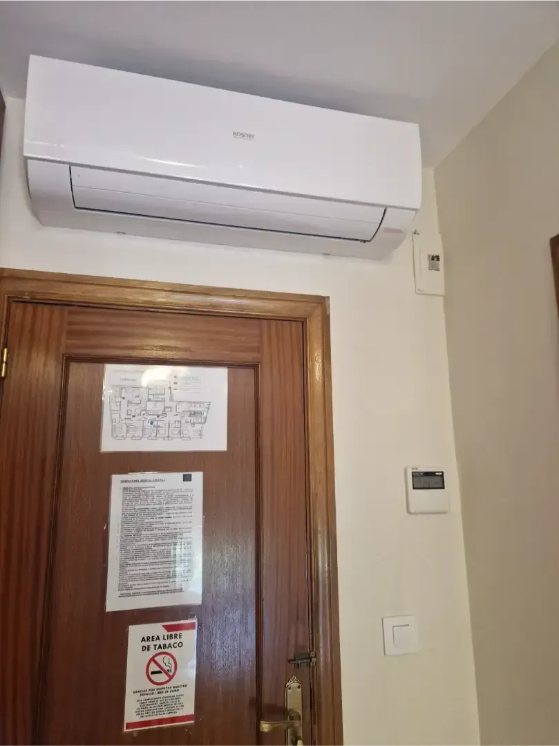 Aire acondicionado blanco montado en la pared, encima de una puerta de madera en una habitación de color beige.