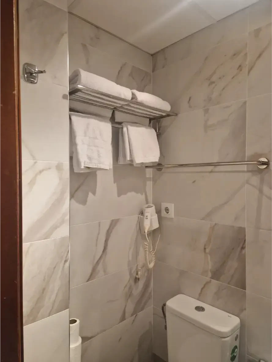 Baño pequeño con azulejos, toallas, secador de pelo de pared, toallero y portarrollos de papel higiénico.