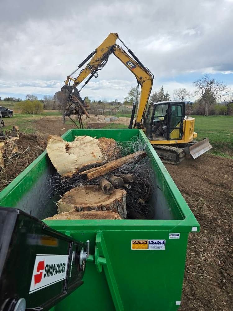 A dumpster filled with logs and a bulldozer in the background.