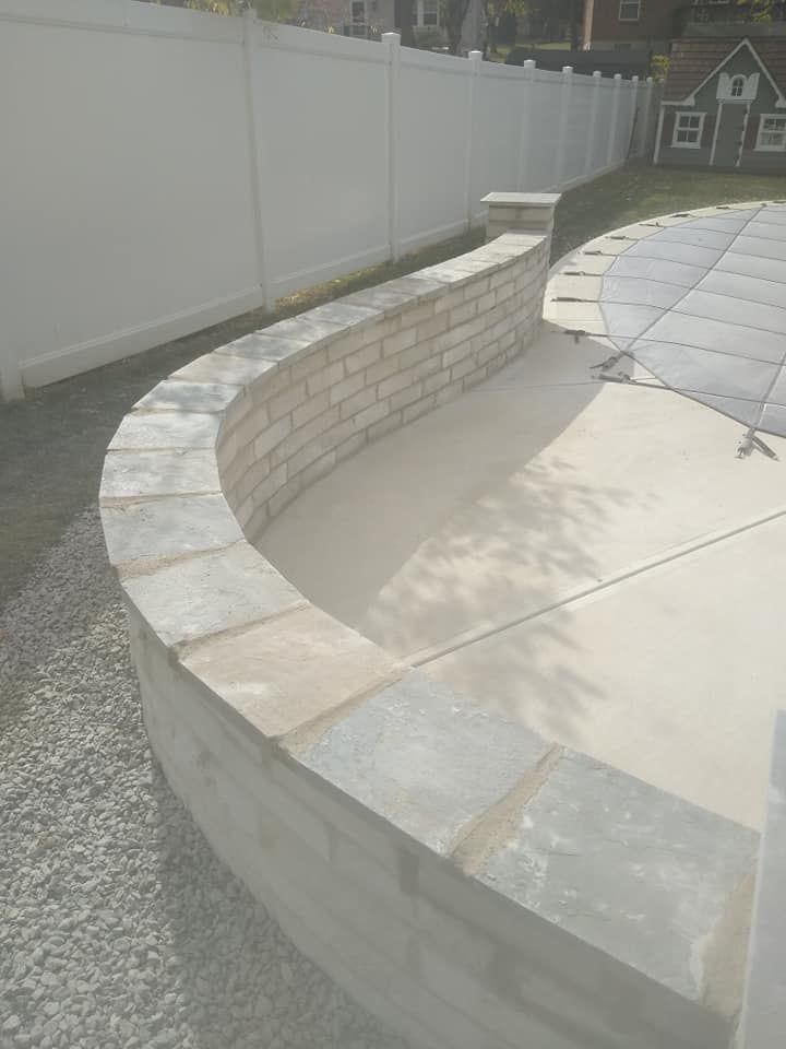 A brick wall is surrounding a pool with a white fence in the background.