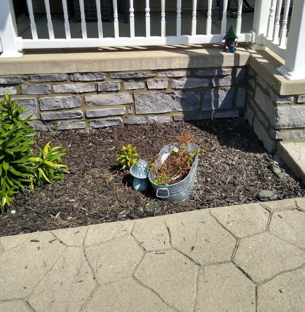A bucket with a plant in it sits in a garden next to a stone wall