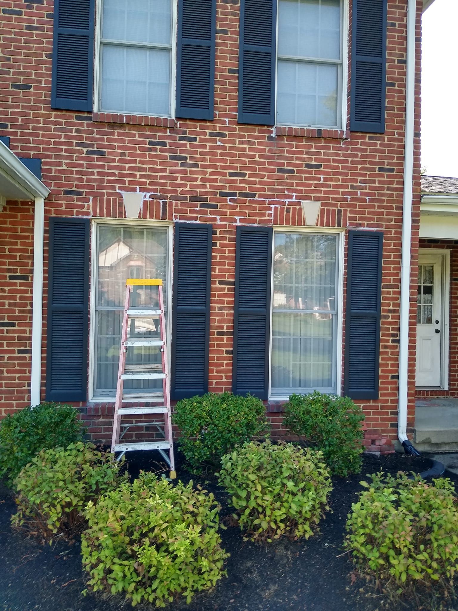 A ladder is sitting in front of a brick house with black shutters
