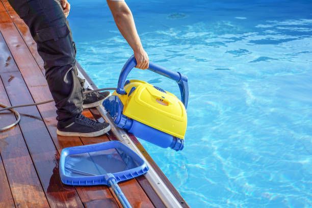 Pool technician on a wooden deck placing an automatic robotic pool cleaner into a bright blue swimming pool.