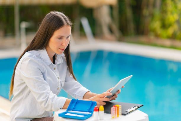 Female pool service professional sitting by a swimming pool checking details on a digital tablet.