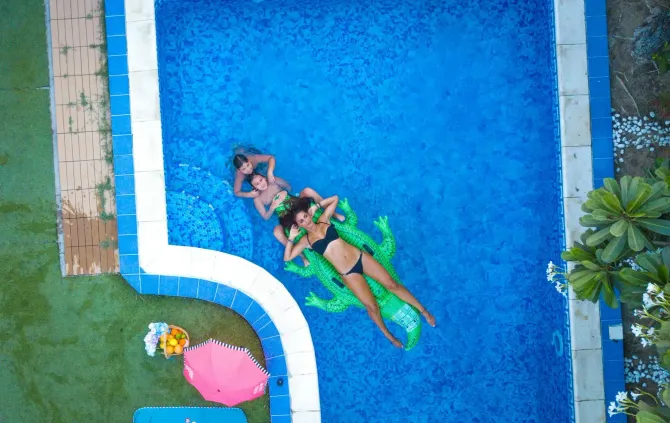 High-angle aerial view of people relaxing on a green inflatable float in a bright blue swimming pool.