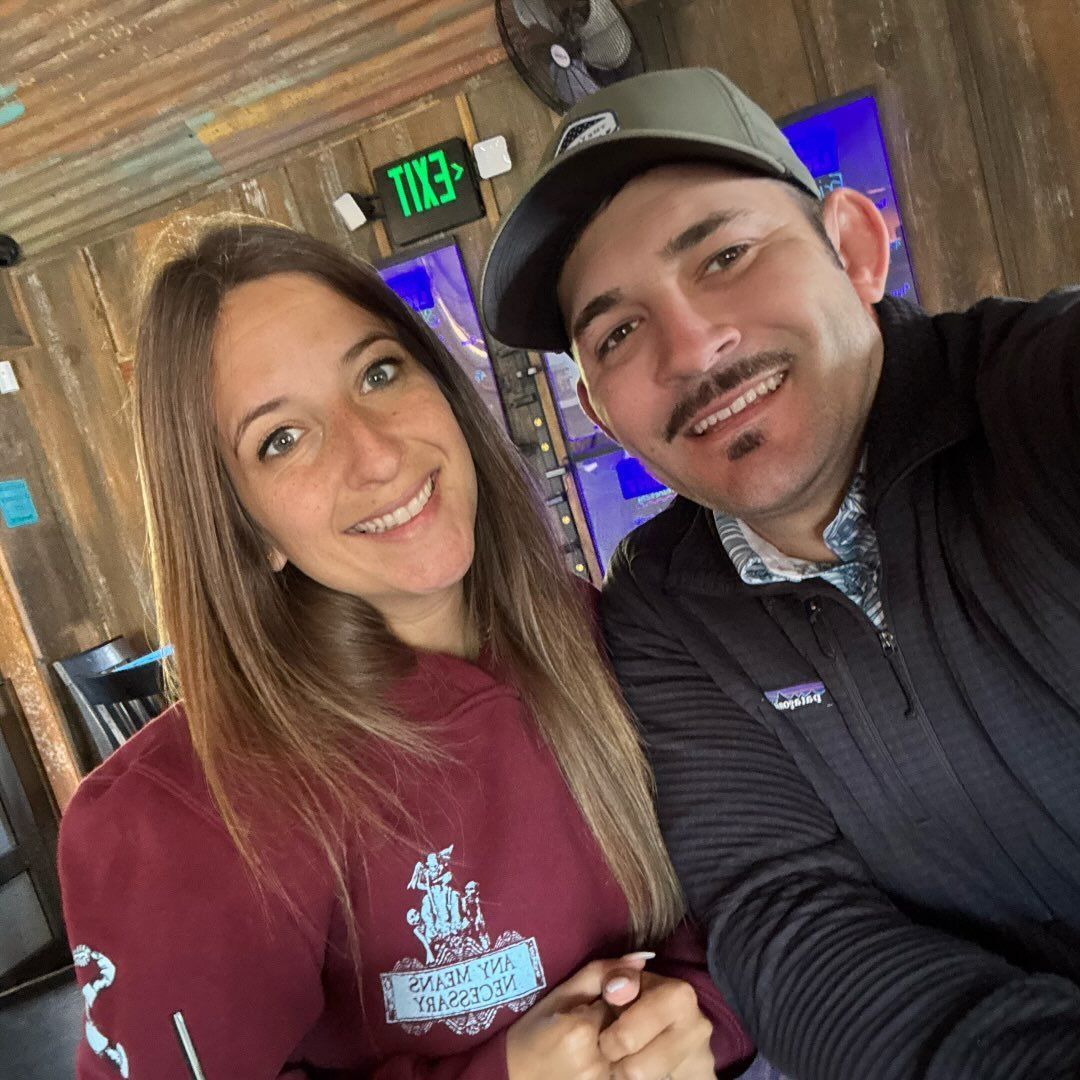 A smiling woman in a maroon hoodie and a man in a cap and dark jacket pose for a selfie indoors, likely at a restaurant.