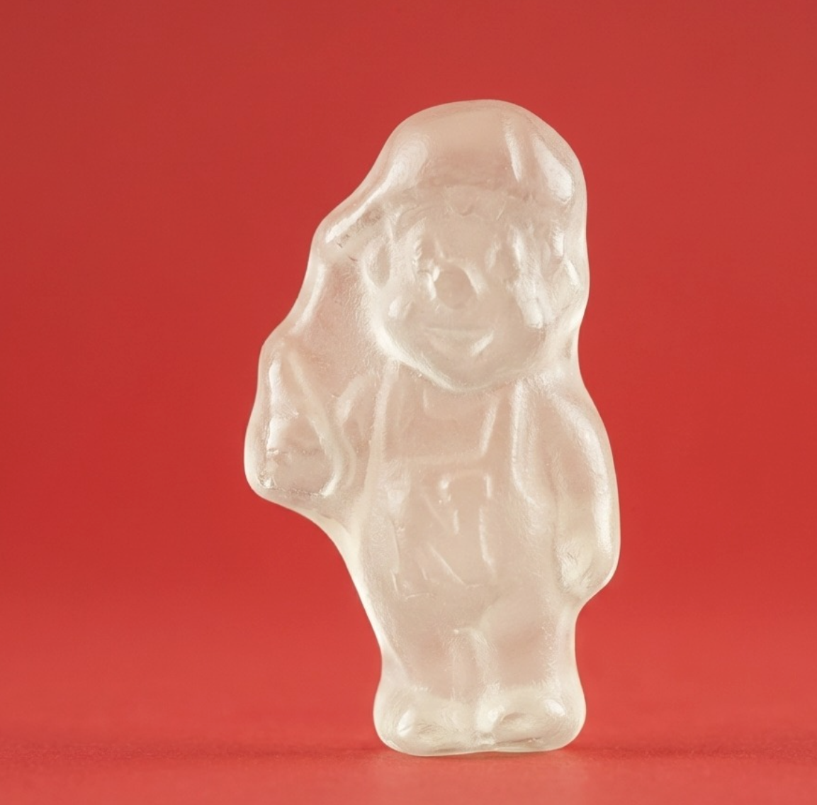 Clear gummy candy shaped like a person with a hat and overalls, on a red background.