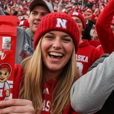 Woman with glasses smiles, resting chin on hand. Wearing denim jacket; brick wall background.