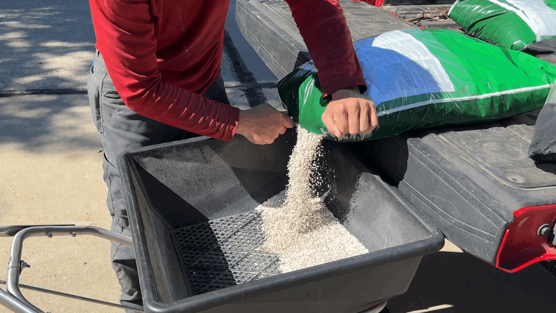 Person pouring grain from a bag into a black seed spreader outdoors