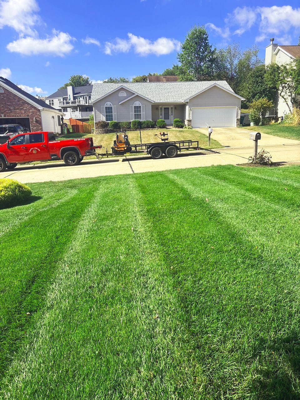 Green lawn with stripes, house, red truck, trailer, blue sky.