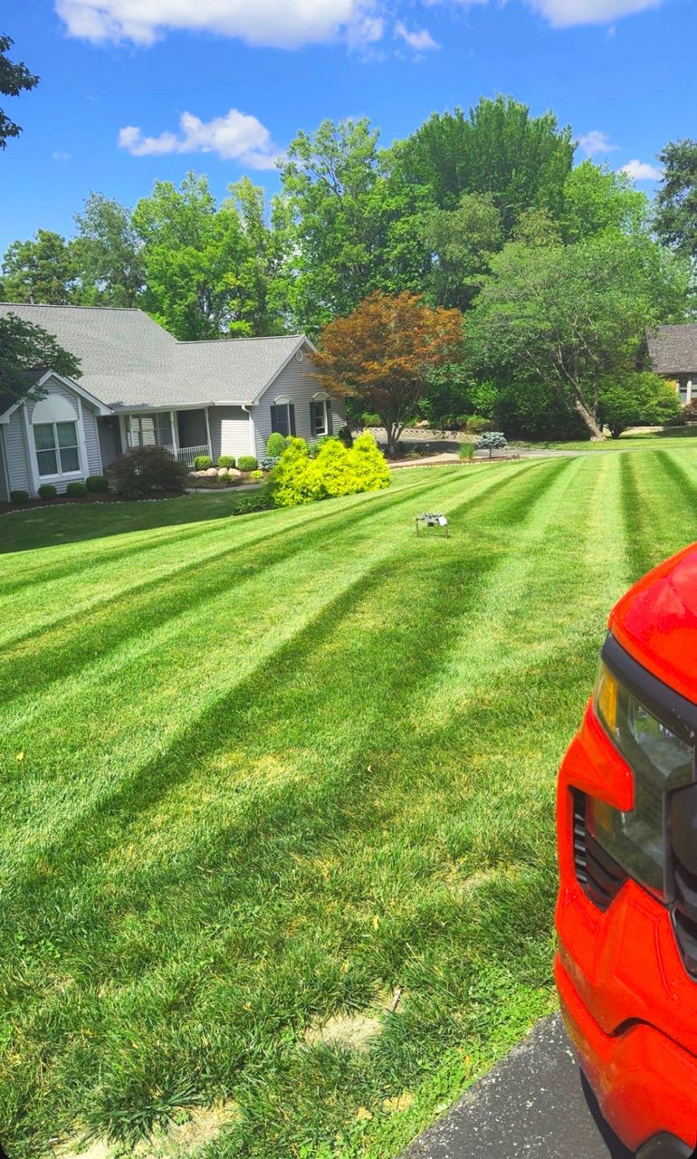 Lawn with freshly cut stripes; red vehicle in the foreground. House and trees in the background on a sunny day.