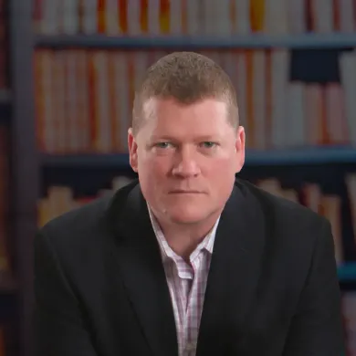 A man in a suit is sitting in front of a bookshelf.