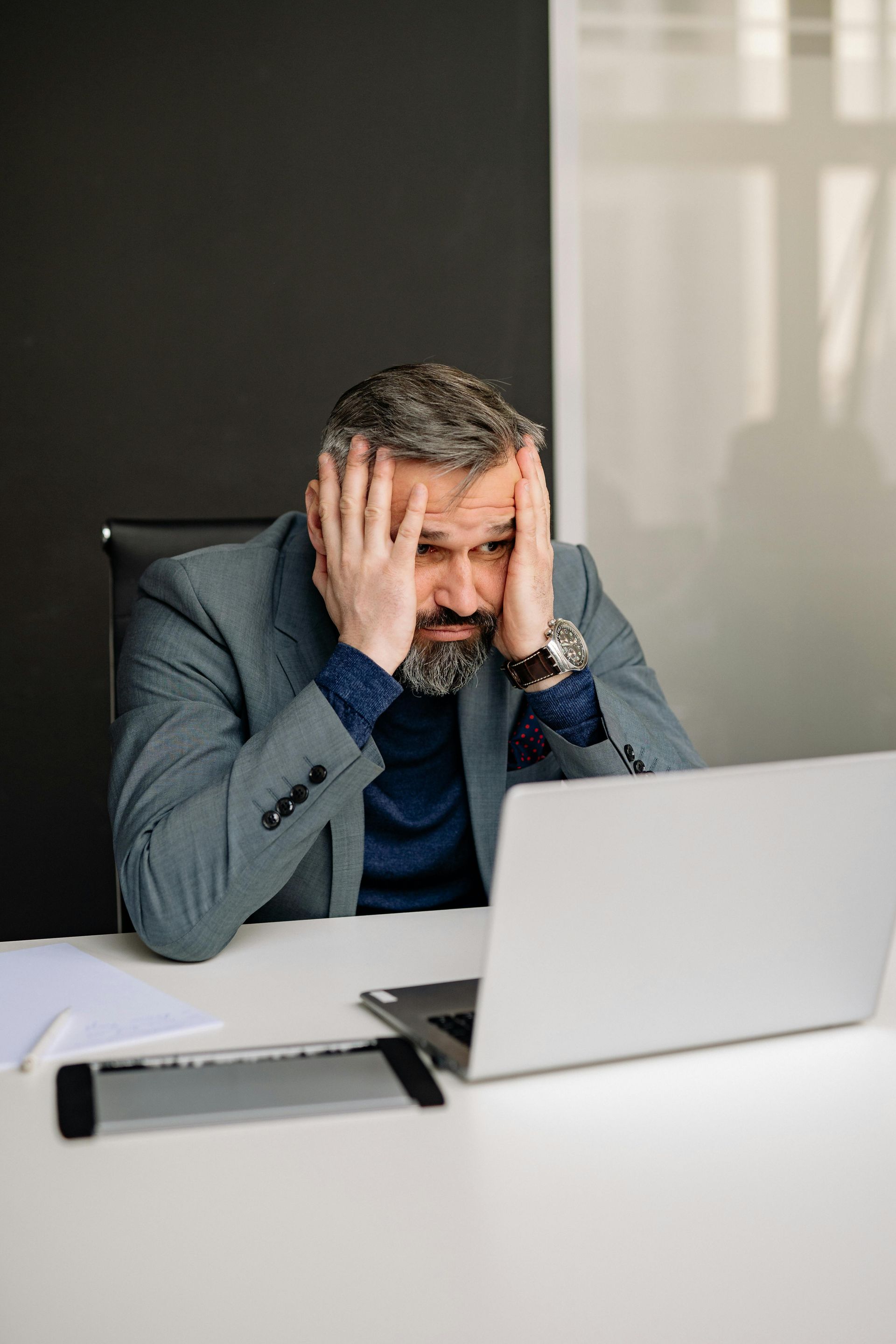 A man is sitting at a desk with his hands on his head in front of a laptop computer.