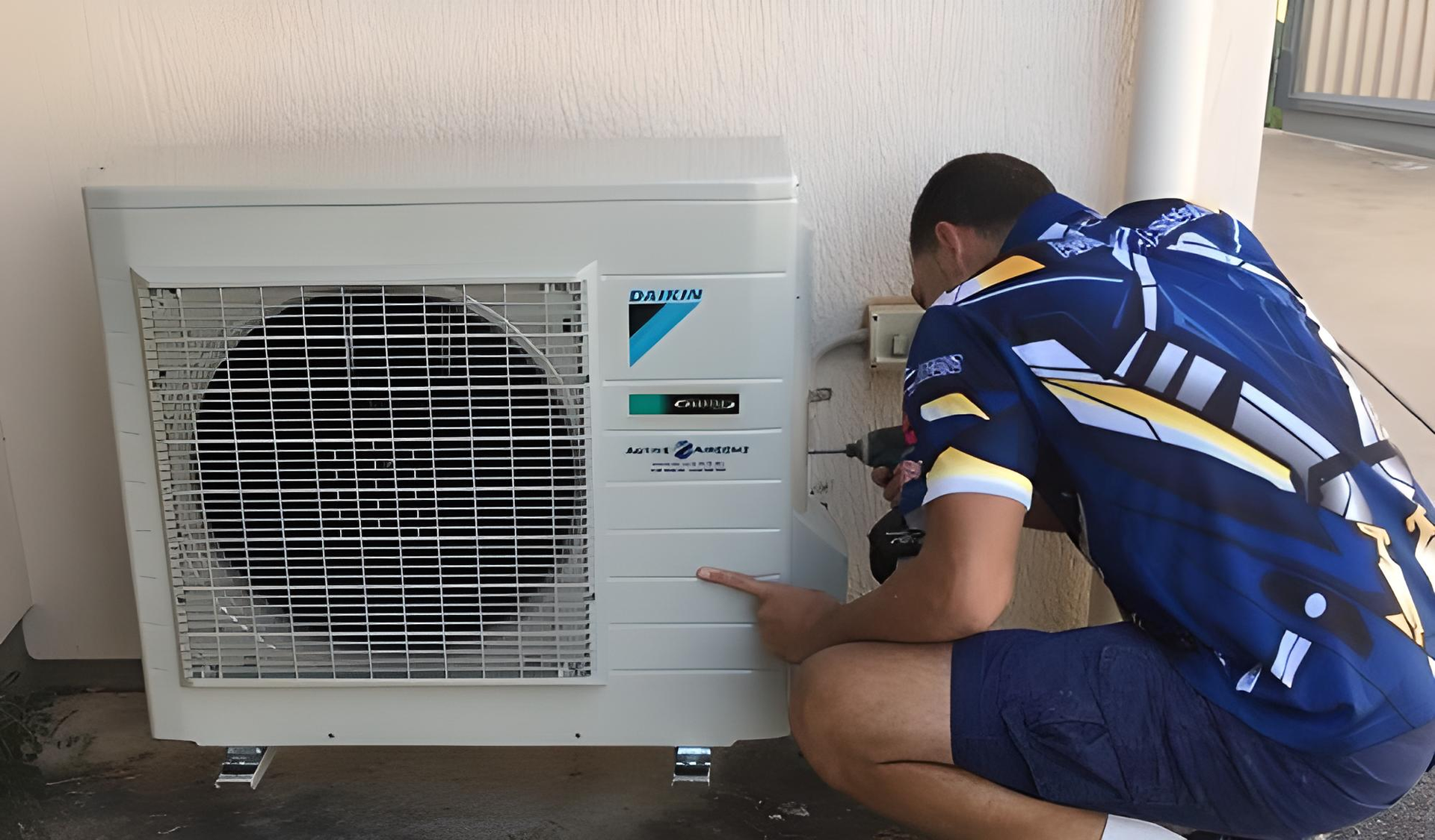 A Technician in A Blue Sports Jersey Uses a Power Drill on The Side of A White Daikin Outdoor Air Conditioning Unit — Active Airconz in Winnellie, NT