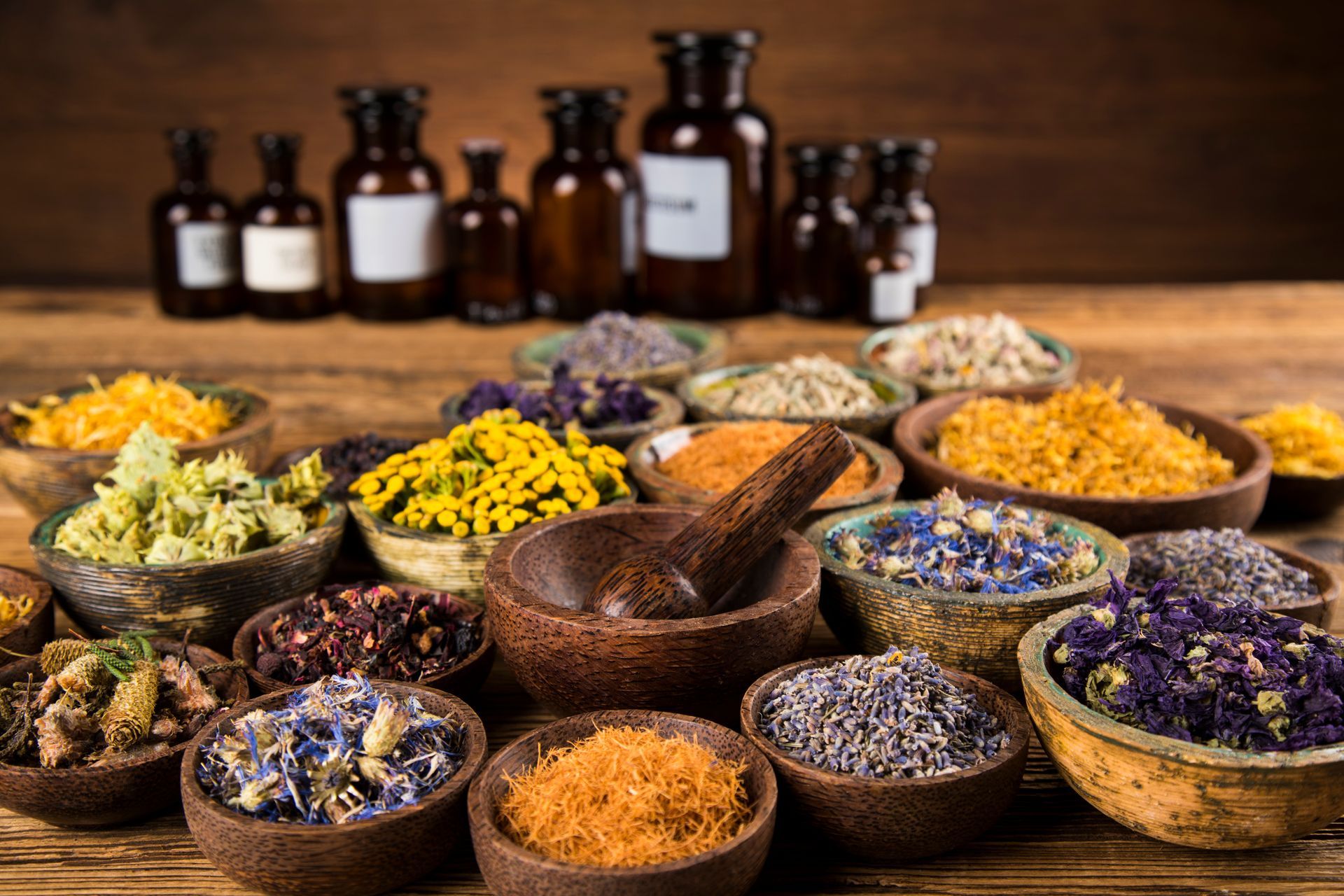 Bowls of dried herbs and flowers, mortar and pestle, apothecary bottles on a wooden table.