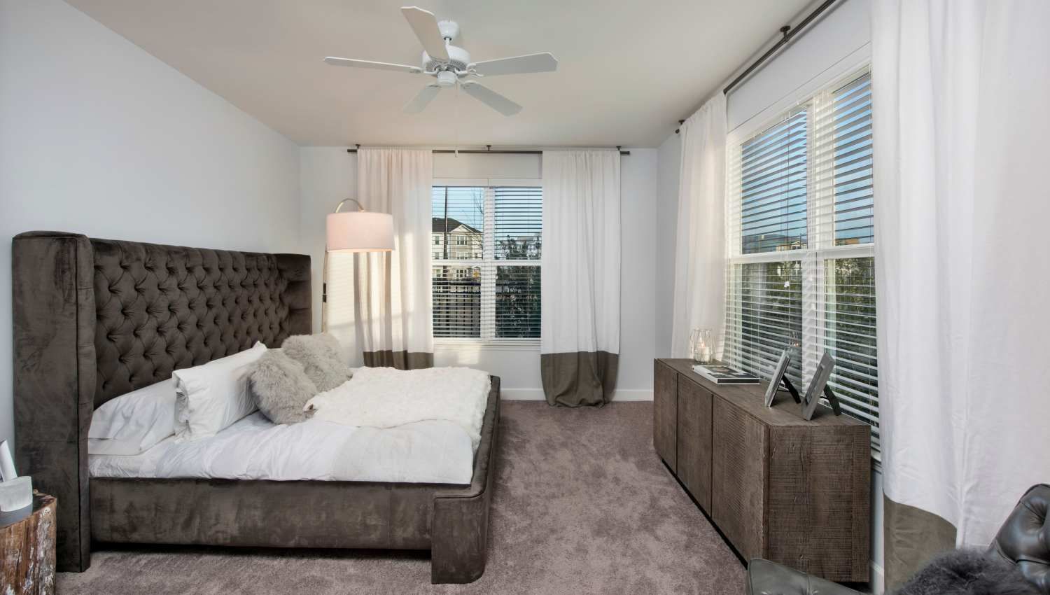 Bedroom with a dark tufted bed, white bedding, and large windows with white curtains.