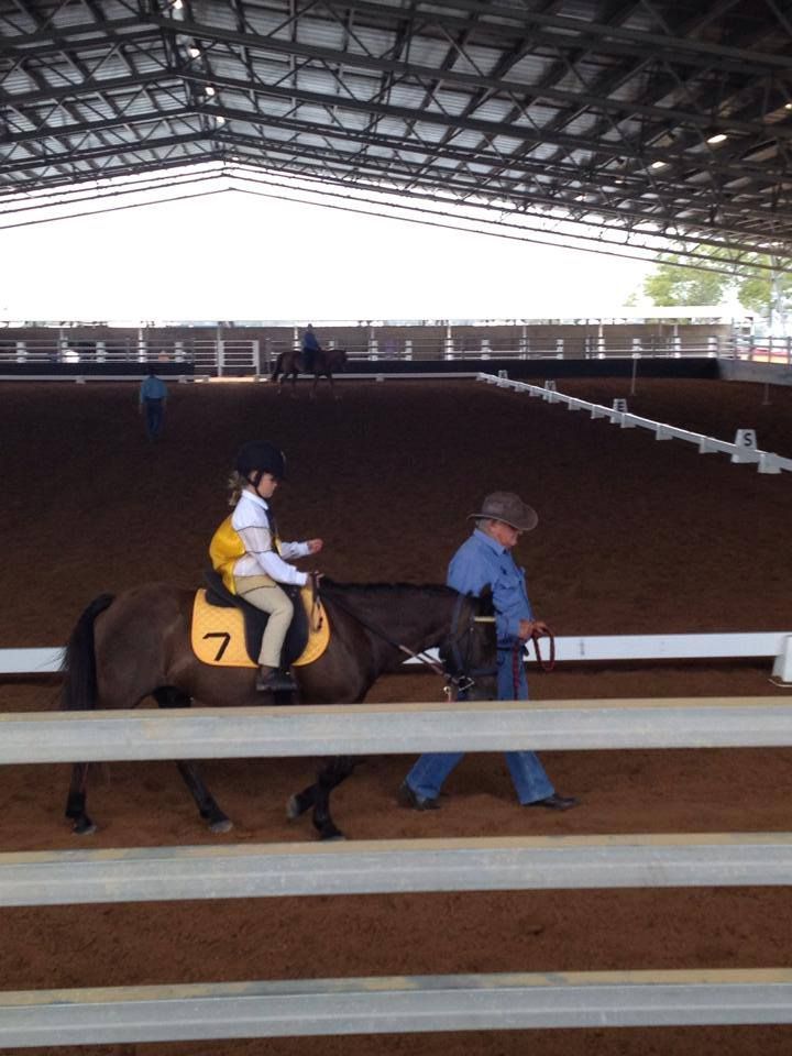 Child riding a horse in arena, being led by an adult. Arena has sand floor and is enclosed.