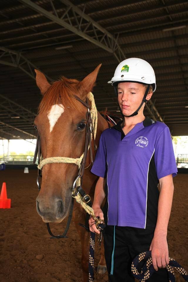 Boy in helmet and purple shirt stands next to a brown horse in an indoor arena.
