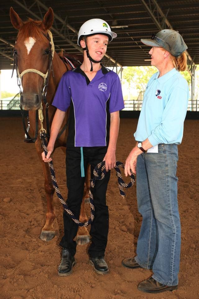 Boy in helmet with horse and instructor in an arena. The boy is talking to the instructor.