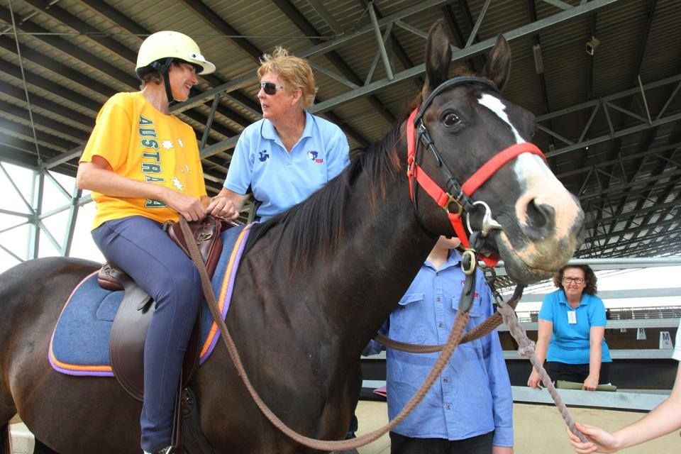 A person in yellow shirt and helmet rides a horse, guided by two helpers under a covered area.
