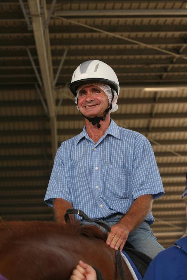 Man wearing a white helmet and blue shirt smiling while sitting on a brown horse.