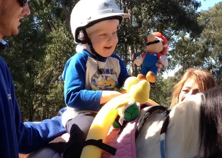 Child on a pony, wearing a helmet, holding Mario plush. Another person helps. Outdoors, sunny.