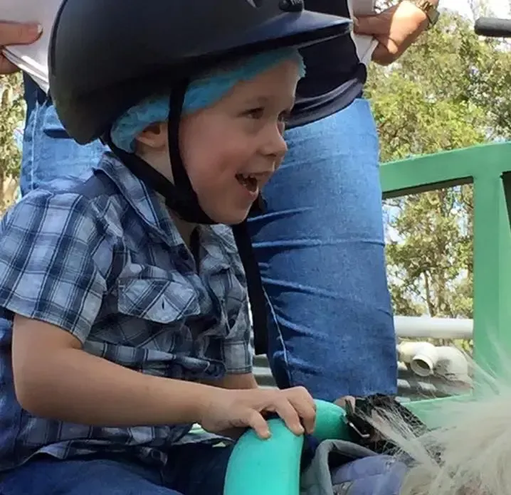 Boy wearing a helmet smiles while holding a turquoise handle; sitting near a person's legs.