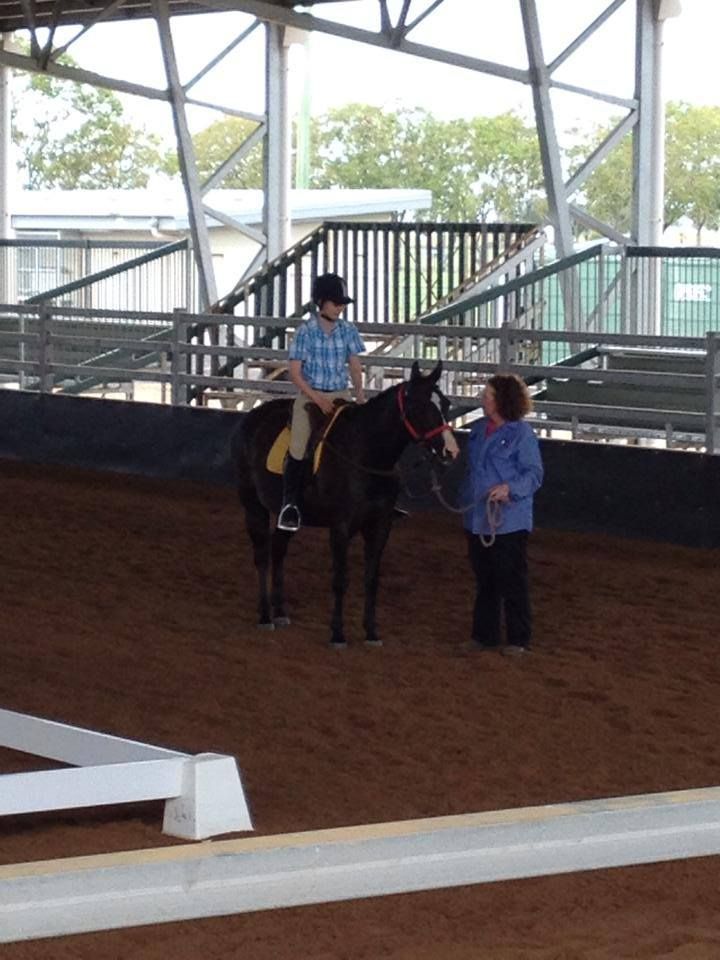 Person on horseback with trainer in an arena. The horse is dark.