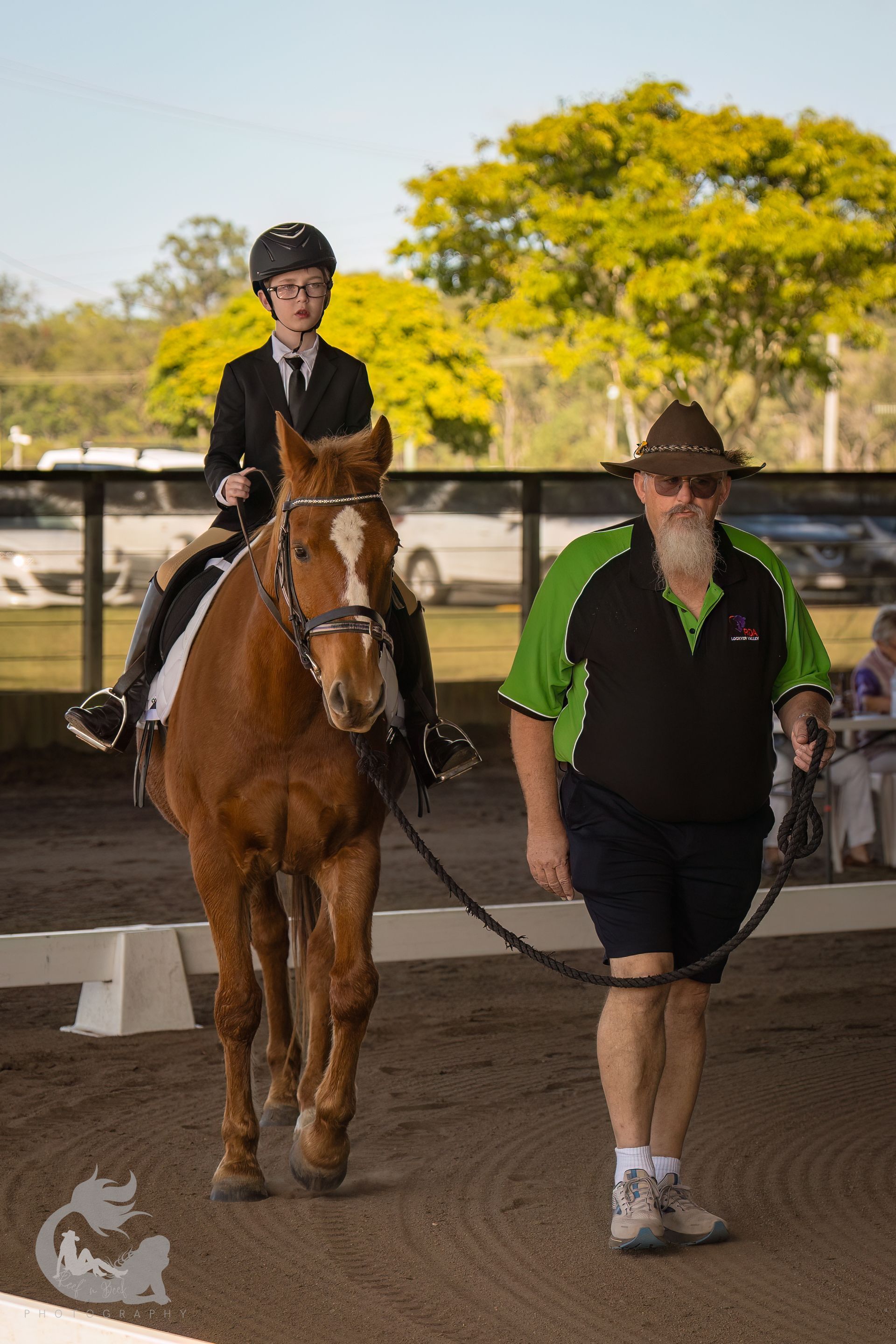 A young rider on a chestnut horse, being led in a dressage arena by a man in a hat.