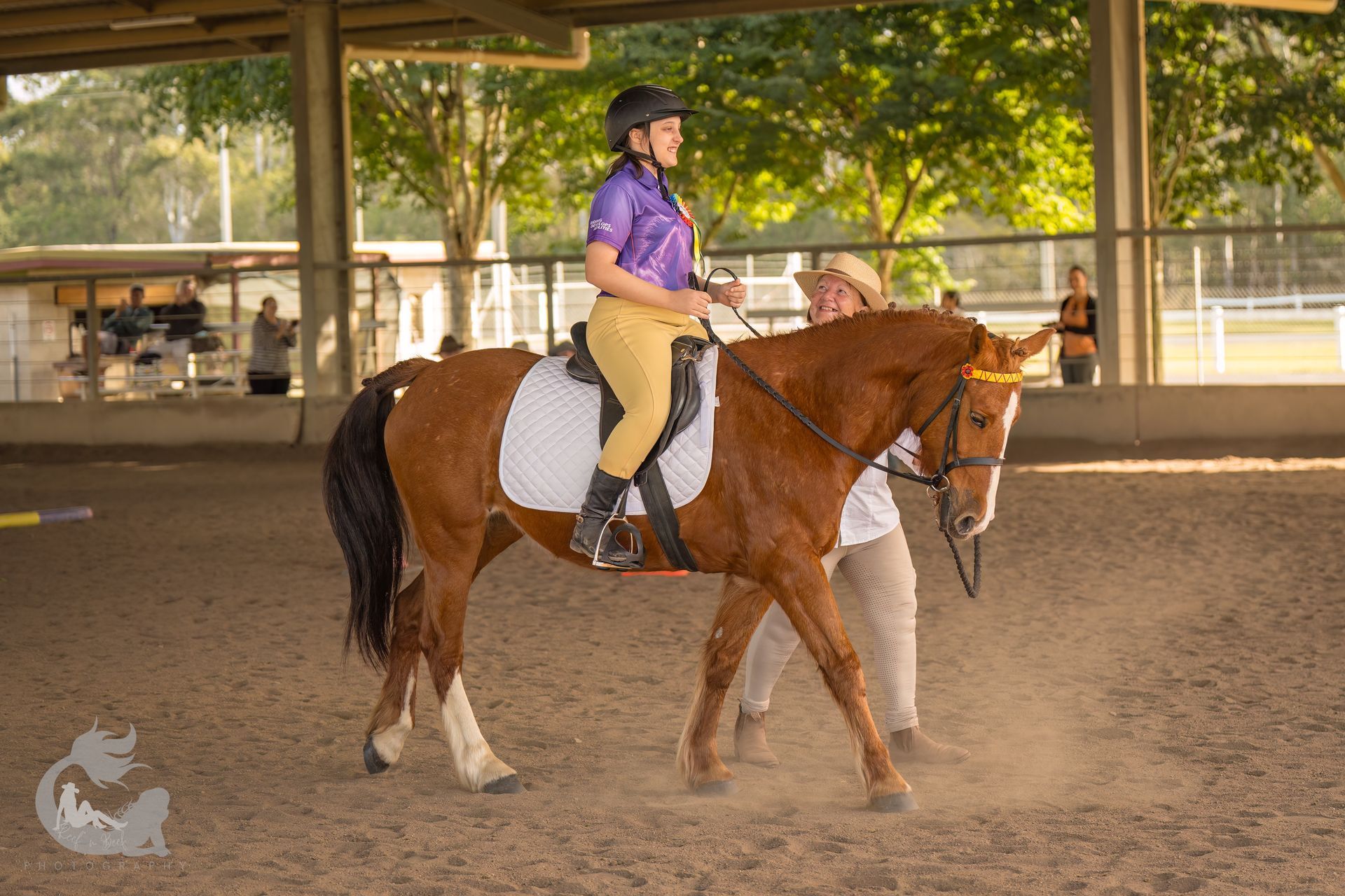Woman on a brown horse in an arena being led by a person. Both wear riding gear.