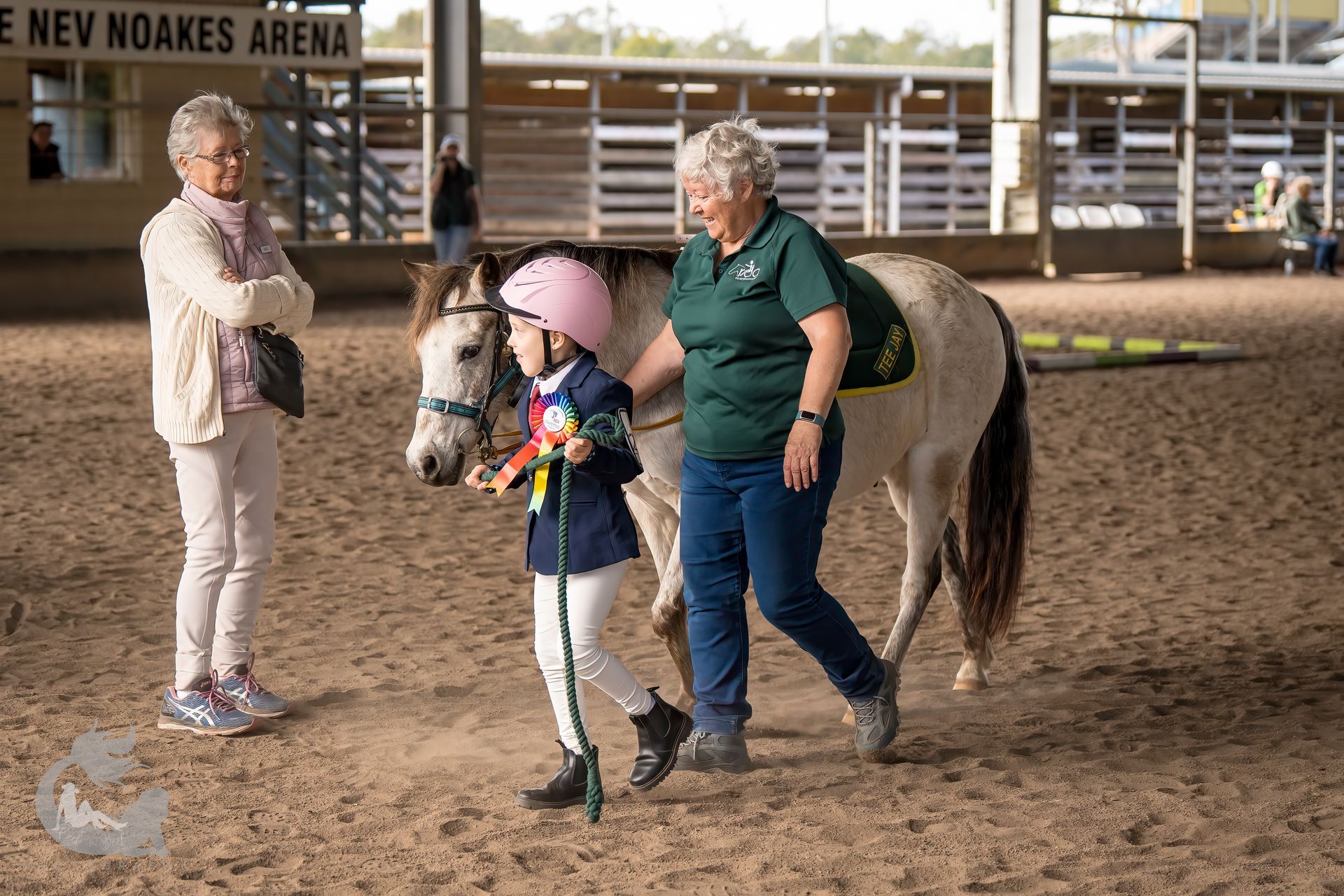 A child in riding gear walks a pony with help from an adult in an indoor arena; another adult observes.
