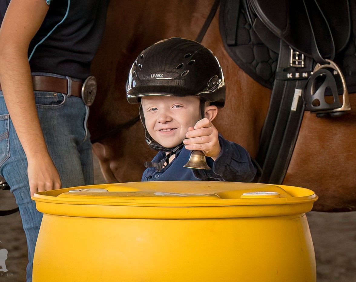 Child in a riding helmet ringing a bell, next to a yellow barrel, near a saddled horse, indoors.
