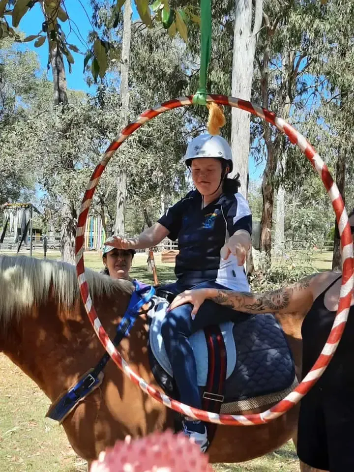 Person on horseback reaching through a hoop with assistance, outdoors.