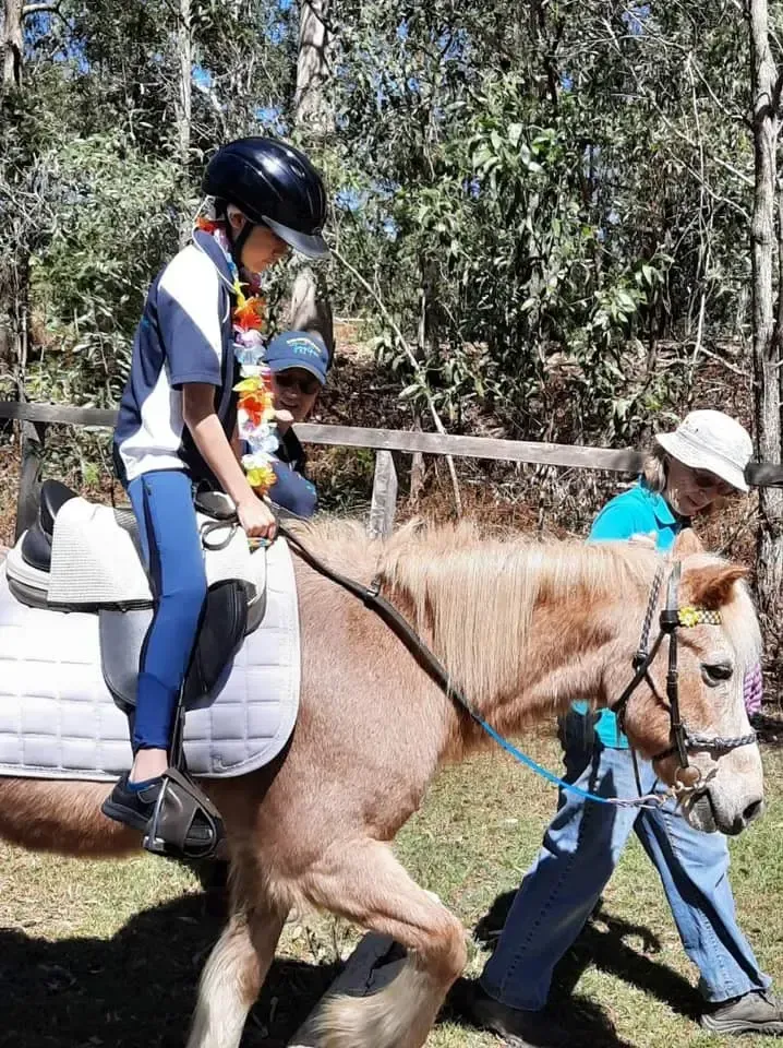 A child in a riding helmet sits on a pony, guided by two adults in an outdoor setting.