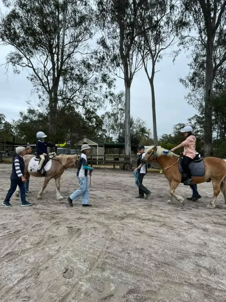 People on horseback in a sandy area; adults lead horses with riders wearing helmets, near trees.