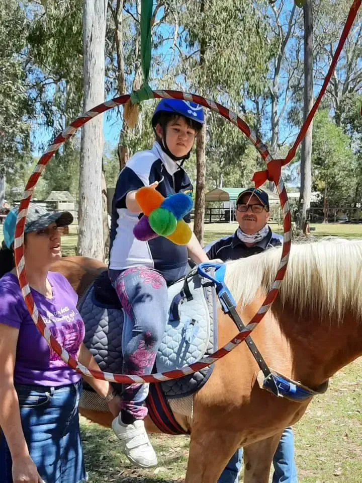 Child riding a horse, holding toy, passing through a hoop with two adults assisting outdoors.