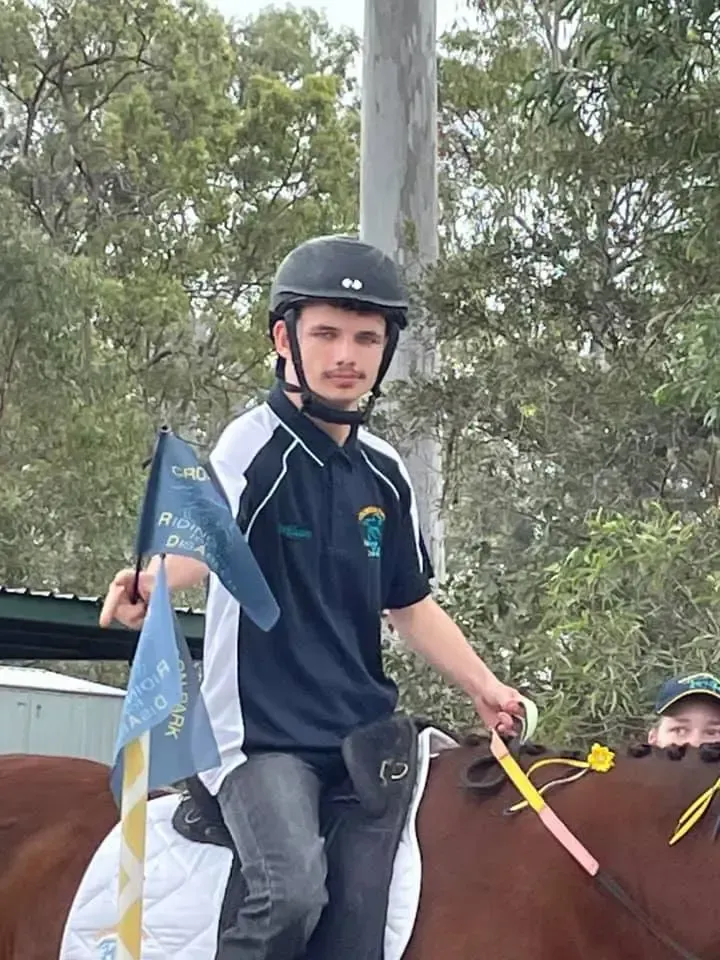 A person on a brown horse, holding a flag, wearing a helmet and navy and white shirt. Outdoors.