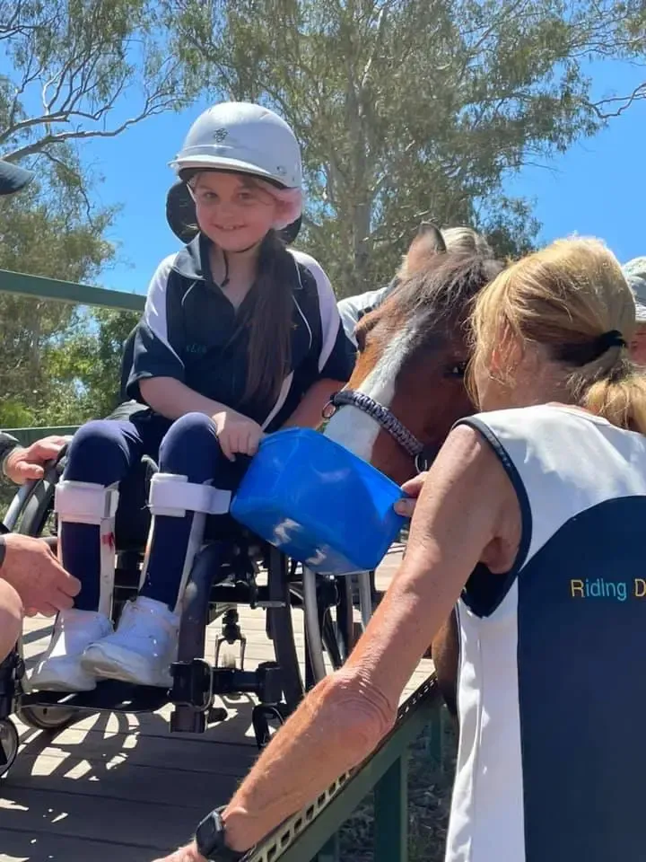 Girl in wheelchair riding a horse, wearing helmet, being assisted by two adults. Blue sky, sunny day.