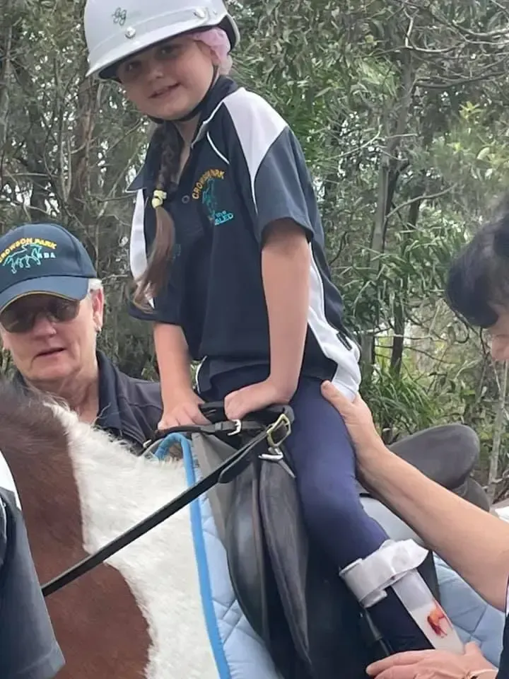 Child riding a horse, wearing helmet, aided by adults. Outdoor setting.