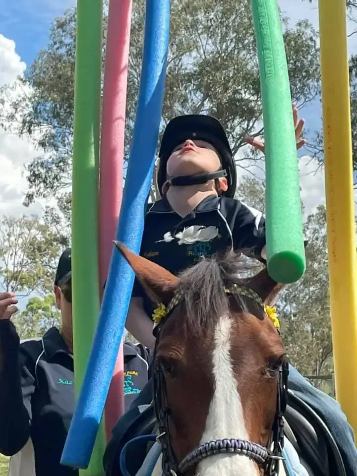 Person on horseback, wearing helmet, reaching up to colorful pool noodles. Outdoor setting.