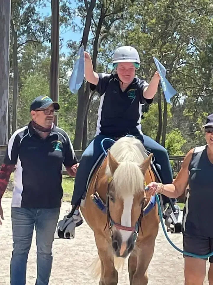 Person riding a horse, holding blue ribbons, smiles. Two people walk alongside. Sunny outdoor setting.