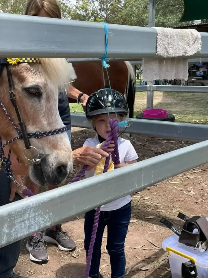 Child in riding helmet holding a purple rope near a pony's head. Woman standing nearby.