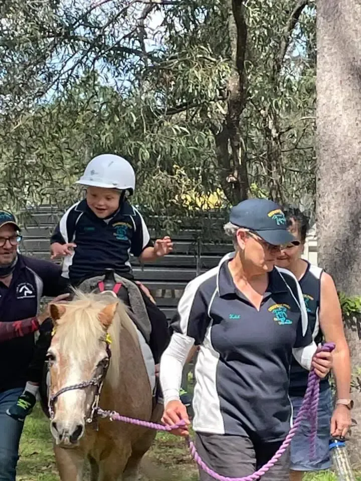 A person in a helmet rides a pony, led by two adults in a park.