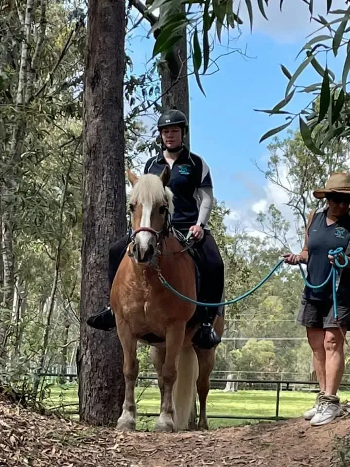 Woman riding a light brown horse in a forest. Another person leads the horse.