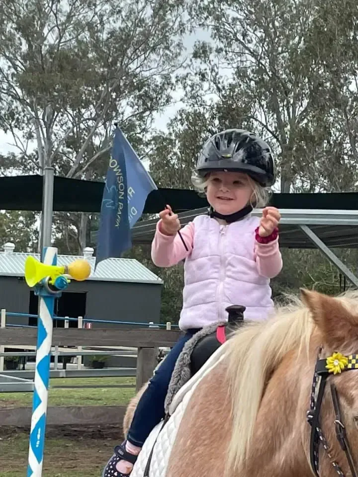 Young child smiles on a pony, wearing a helmet and pink vest, outdoors.