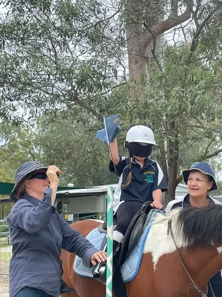 Child on horseback waving, wearing helmet, aided by two adults outdoors.